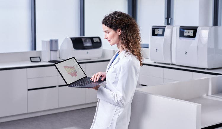 Dental professional reviewing a digital restoration design on a laptop in a manufacturing workspace with milling units.